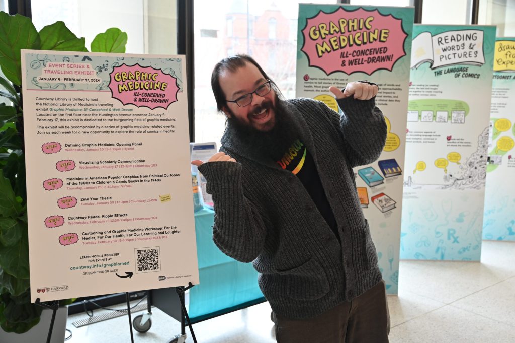Matthew Noe, smiling and gesturing in a funny way towards a poster describing graphic medicine events